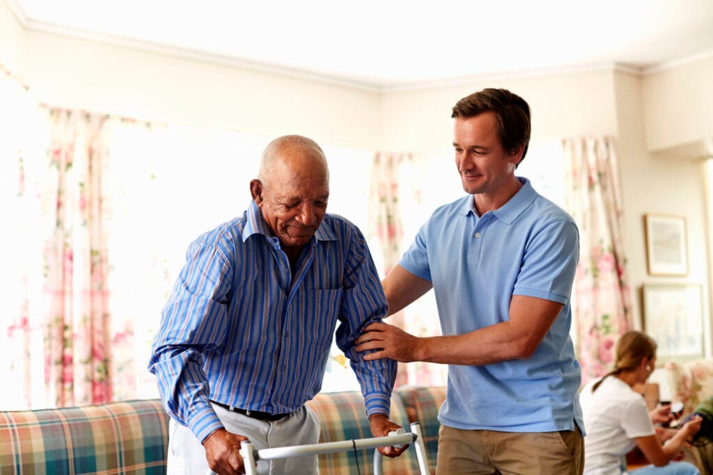 *   Image content: An elderly Black man using a walker, being assisted by a younger white man in a blue polo shirt. They are