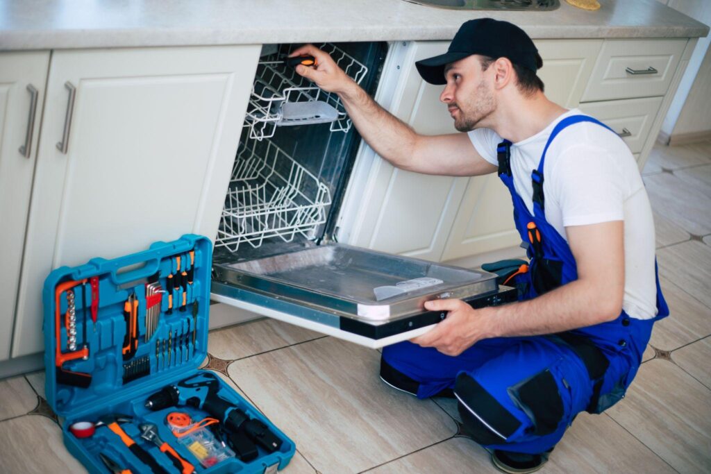 *   Image: A man in blue overalls and a black cap is repairing a dishwasher in a kitchen.
    *   Details: He's kneeling, hol