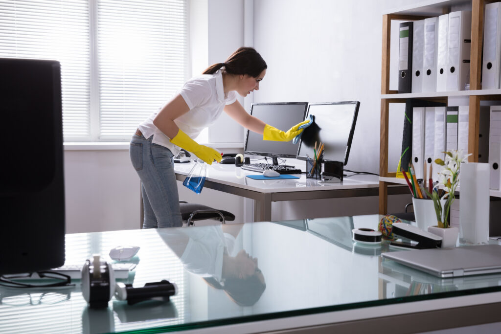 Woman wearing yellow gloves cleaning a computer monitor in a modern office.