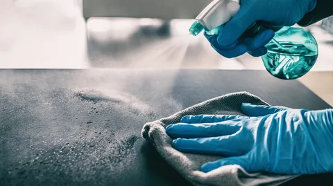 Gloved hands spraying cleaning fluid on a dark surface and wiping it with a cloth.