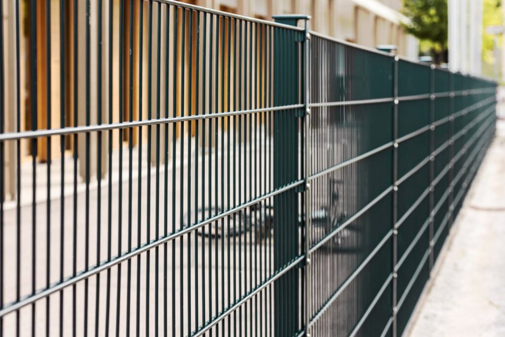 Long dark green metal wire fence with vertical bars running alongside a building.