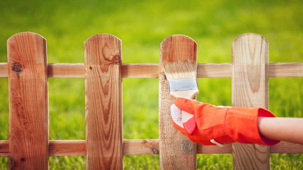 Hand in an orange glove using a paintbrush to apply stain to a wooden picket fence.