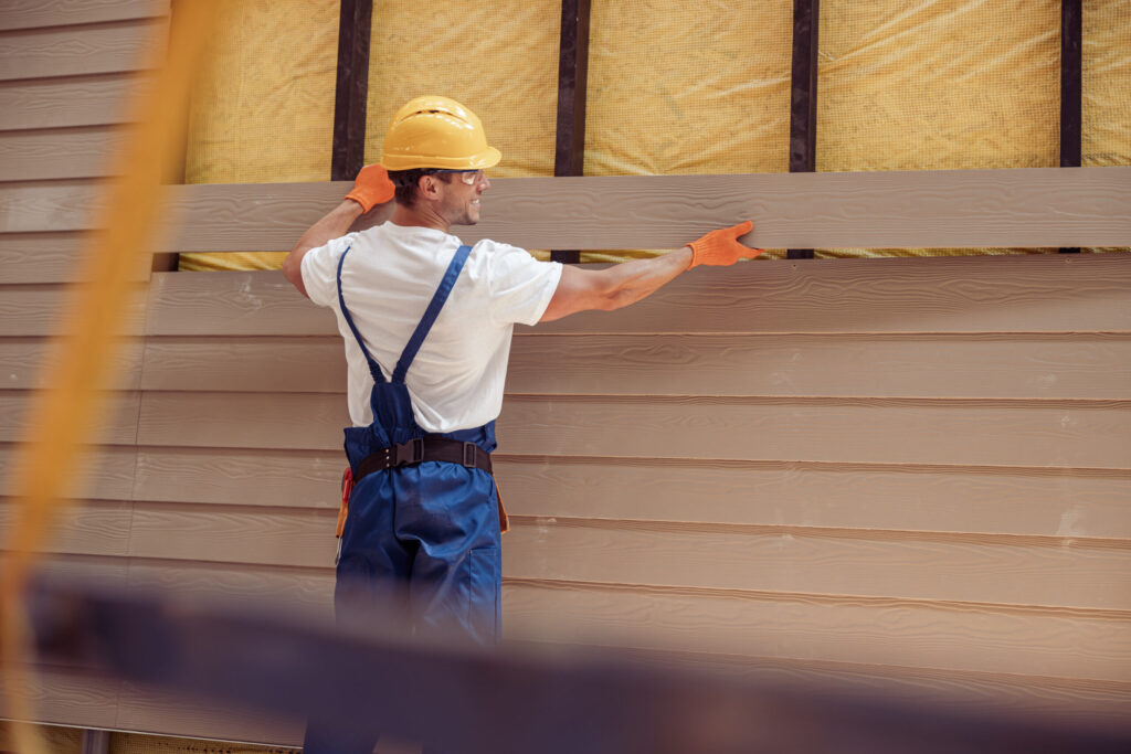 Construction worker in a hard hat and overalls installing tan siding on a building exterior.