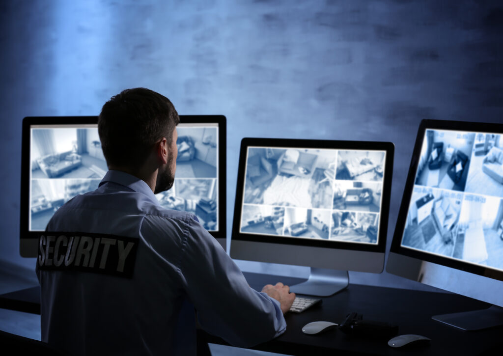 Security guard monitoring surveillance feeds on three computer monitors in a dimly lit room.
