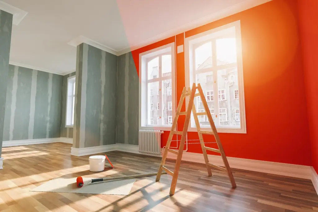 Empty room under renovation with a wall partly painted orange, a stepladder, paint roller, and bucket on wooden floors.
