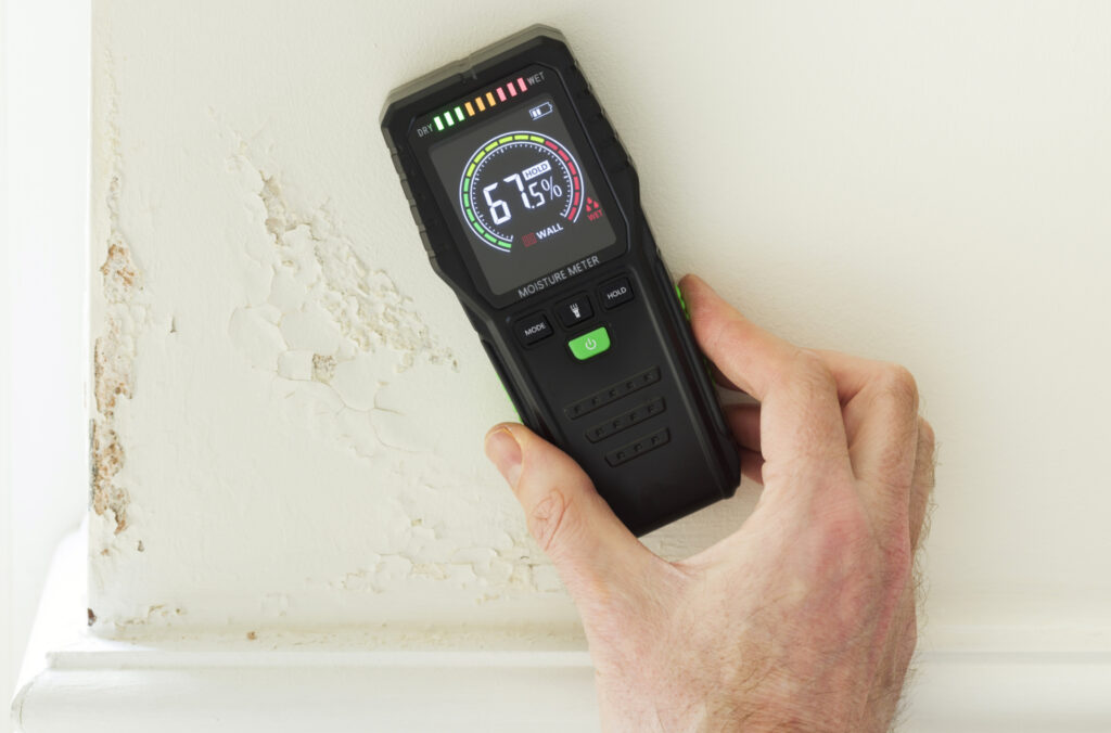 Digital moisture meter showing 67.5% against a white wall with peeling, water-damaged paint.
