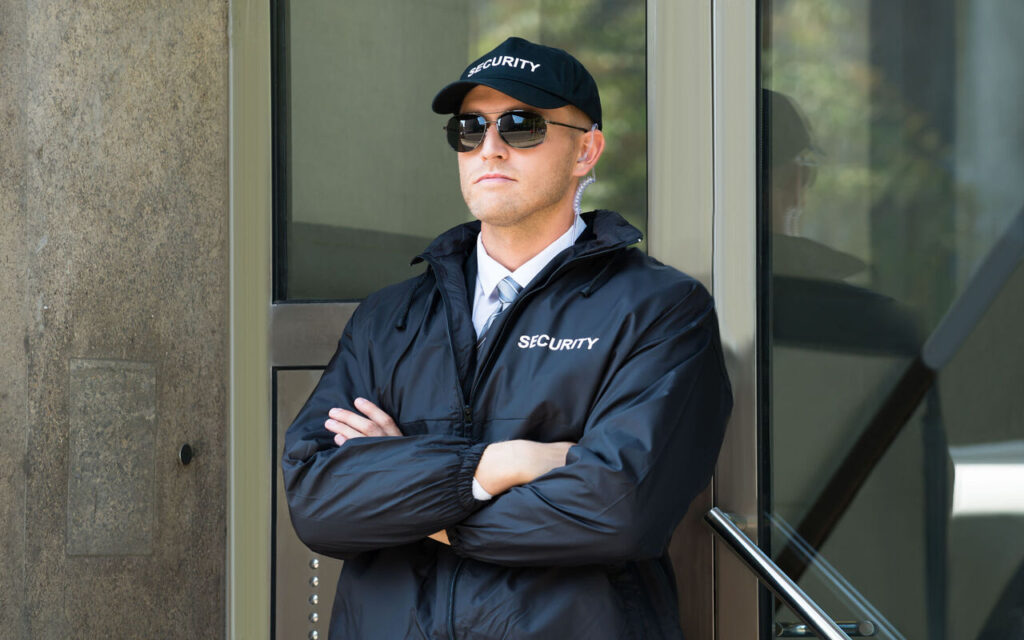 Security guard in a black jacket, cap, and sunglasses standing with arms crossed at a building entrance.