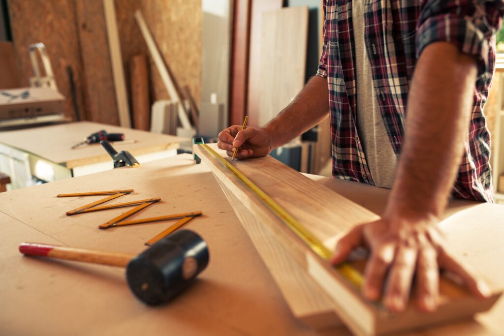 Carpenter marking a piece of wood with a pencil and measuring tape in a workshop.