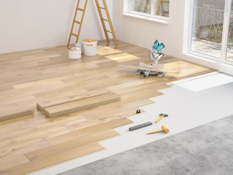 Wooden floor being installed over a concrete subfloor with a miter saw, ladder, and tools scattered nearby.