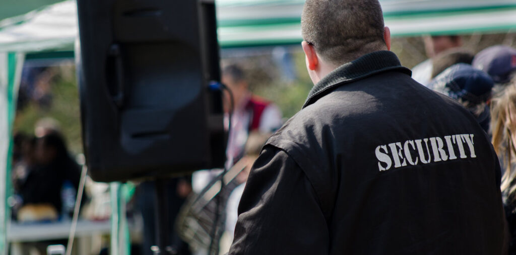 Security guard in a black jacket with "SECURITY" on the back, standing by a large speaker at an outdoor event.