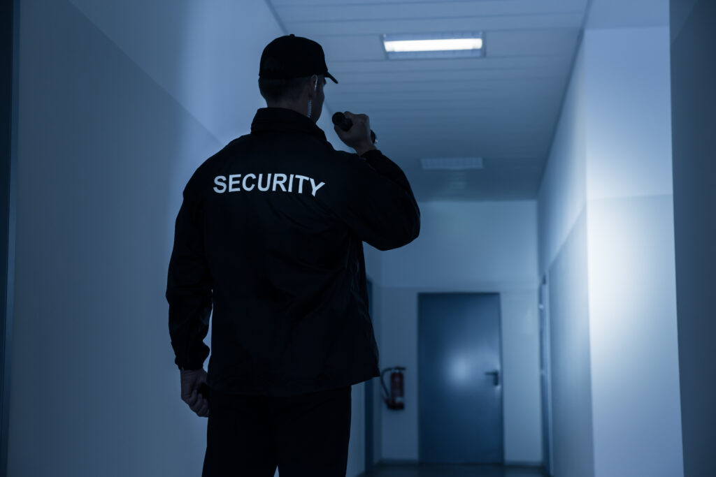 Security guard from behind in a black uniform, holding a flashlight in a dimly lit blue hallway.