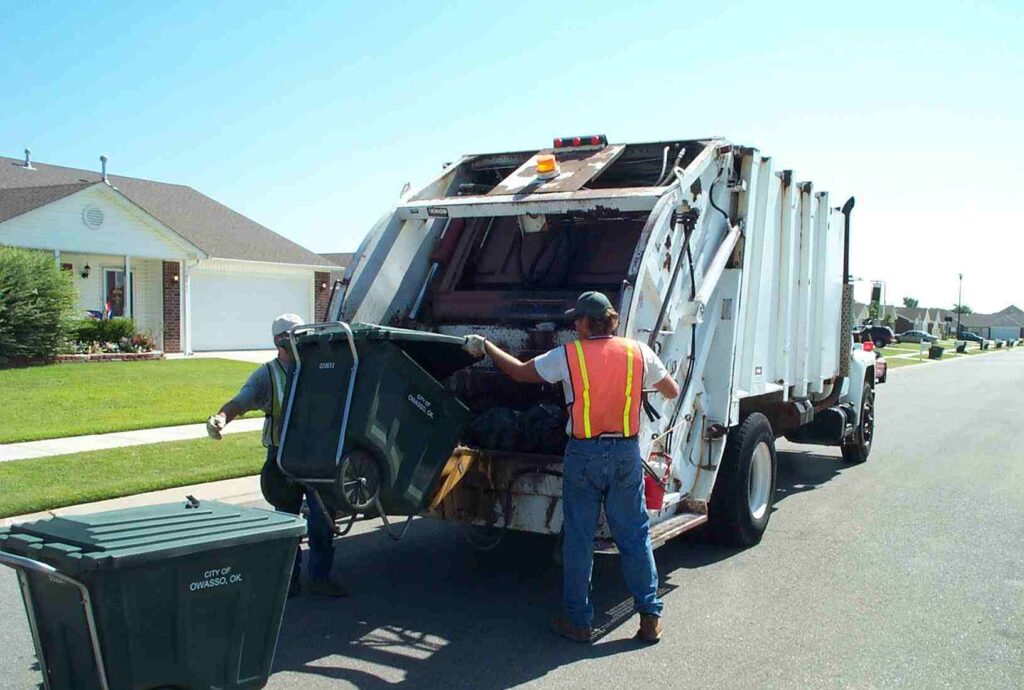 Two sanitation workers empty a green trash bin into the back of a large white garbage truck on a residential street.