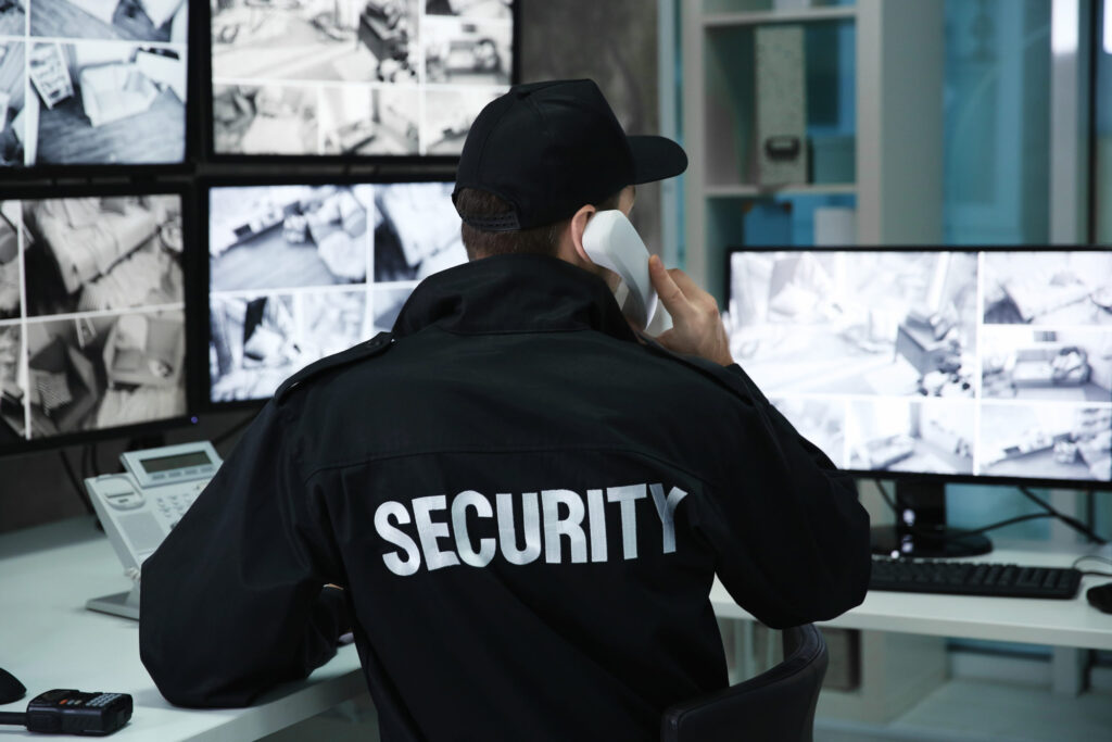 Security guard in black uniform talking on a phone while monitoring CCTV surveillance screens.