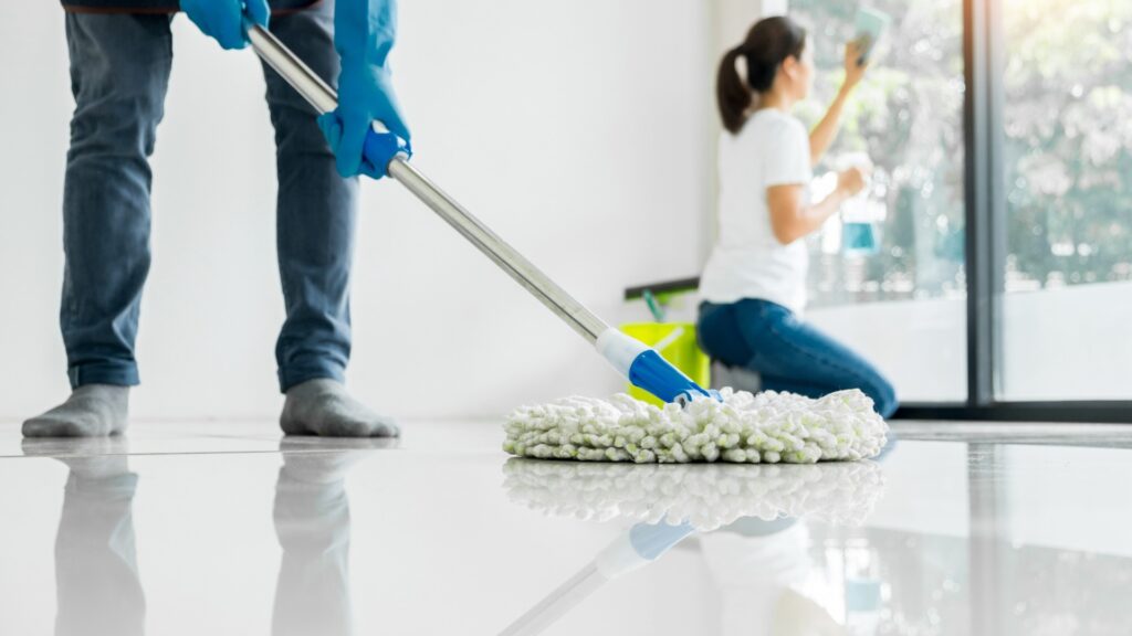 A person mopping a white tiled floor while another person cleans a window in the background.