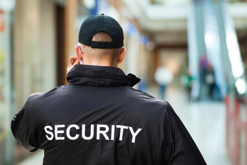 Security guard from behind, wearing a black uniform and cap, adjusting an earpiece.
