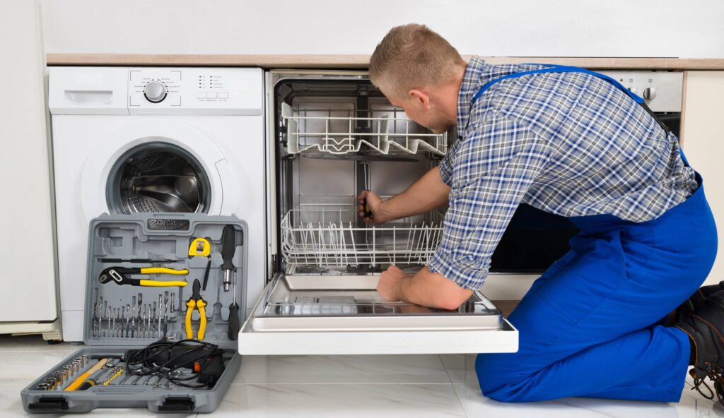 Repairman in blue overalls fixing an open dishwasher with a toolkit on the floor.