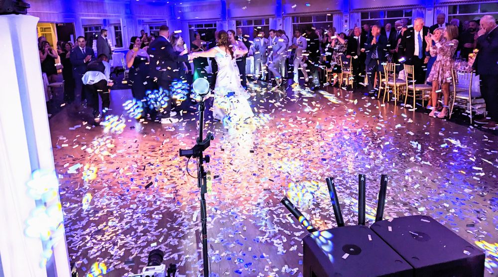 A couple dances on a confetti-covered dance floor in a ballroom filled with guests and stage lighting.