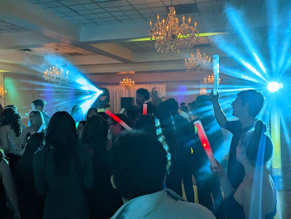 Crowd dancing under chandeliers in a ballroom with stage lighting and glowing foam sticks.