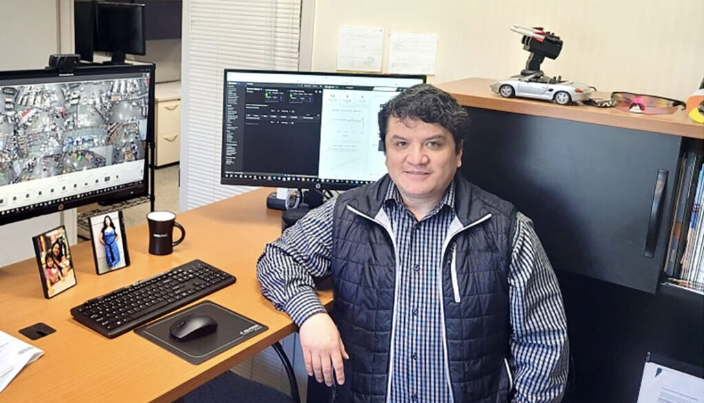 A man in a black vest sits at a desk with two computer monitors, a keyboard, a mouse, and a mug.