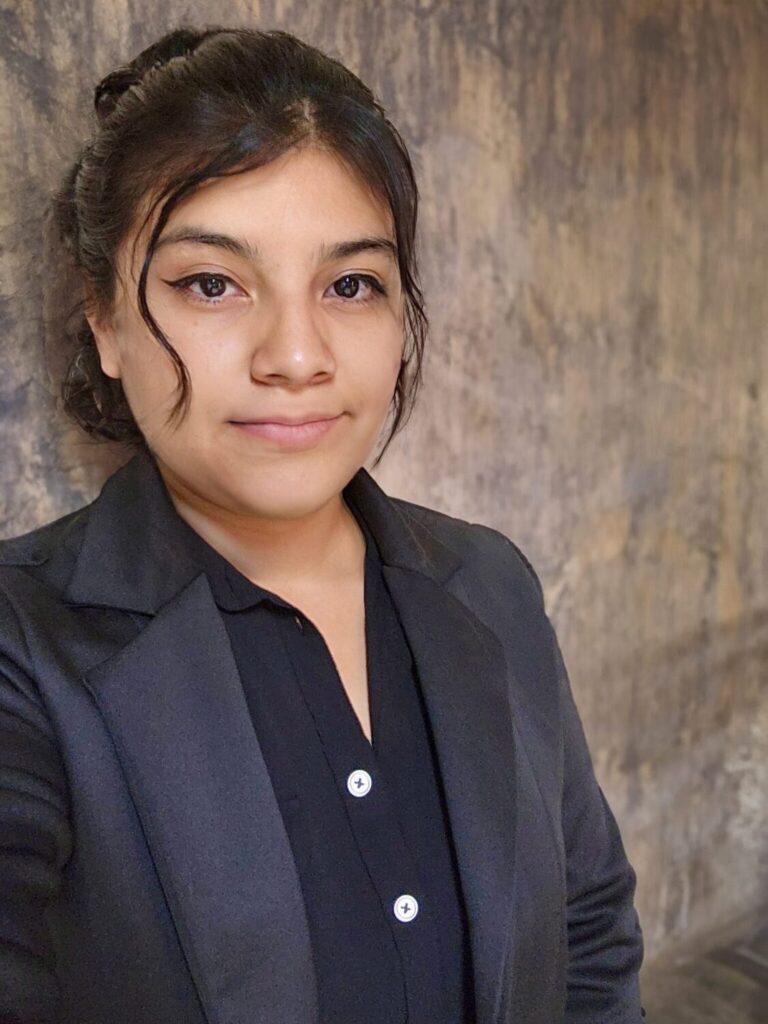 Headshot of a woman with dark hair in a bun, wearing a black blazer and button-down shirt against a textured wall.