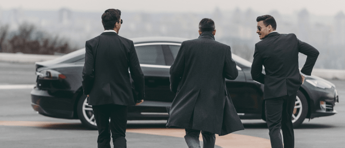 Three men in black suits seen from behind, standing in front of a black luxury car in a foggy outdoor setting.