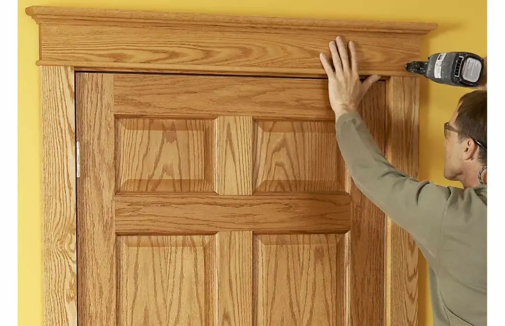 A person uses a power nailer to attach oak trim to the top of a door frame.