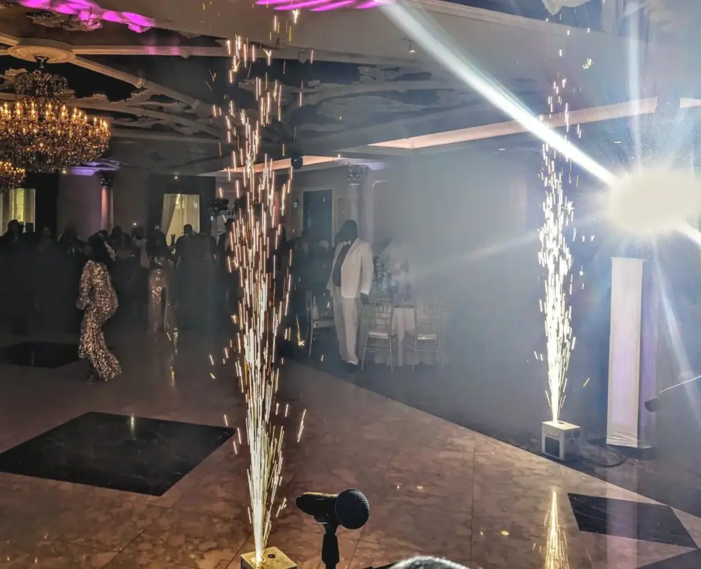 Spark fountains erupt in a dim ballroom during a celebration with guests dancing and a man in a white suit.