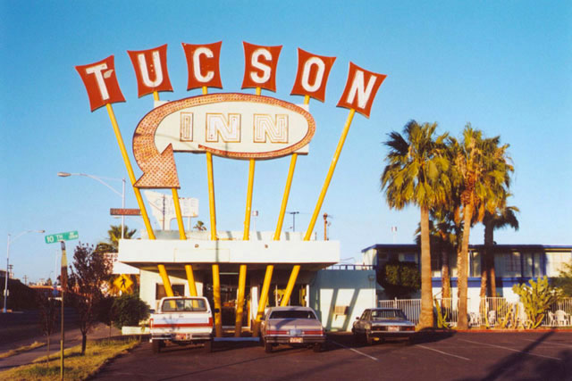 A large, vintage "Tucson Inn" neon sign stands over the motel entrance in a parking lot with palm trees under a blue sky.