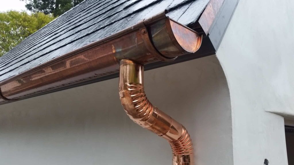 Polished copper gutters and curved downspout on a white wall under a dark slate roof.