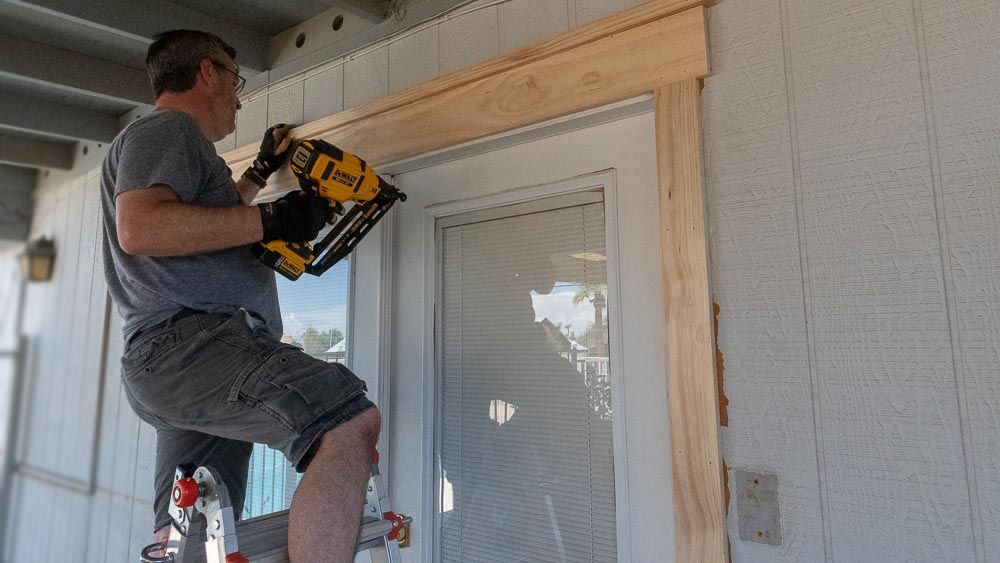 A man on a ladder uses a nail gun to attach wooden trim around an exterior door.