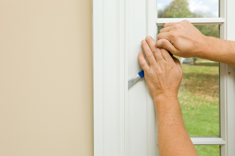 Hands using a putty knife to remove trim from a window frame.