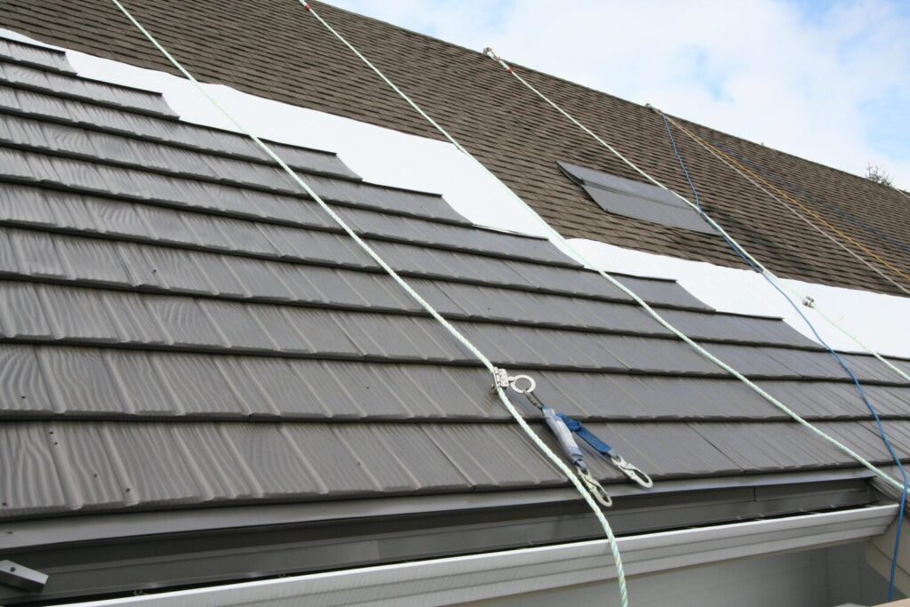 Grey metal roofing tiles being installed over white underlayment with safety lines and fall protection clips.