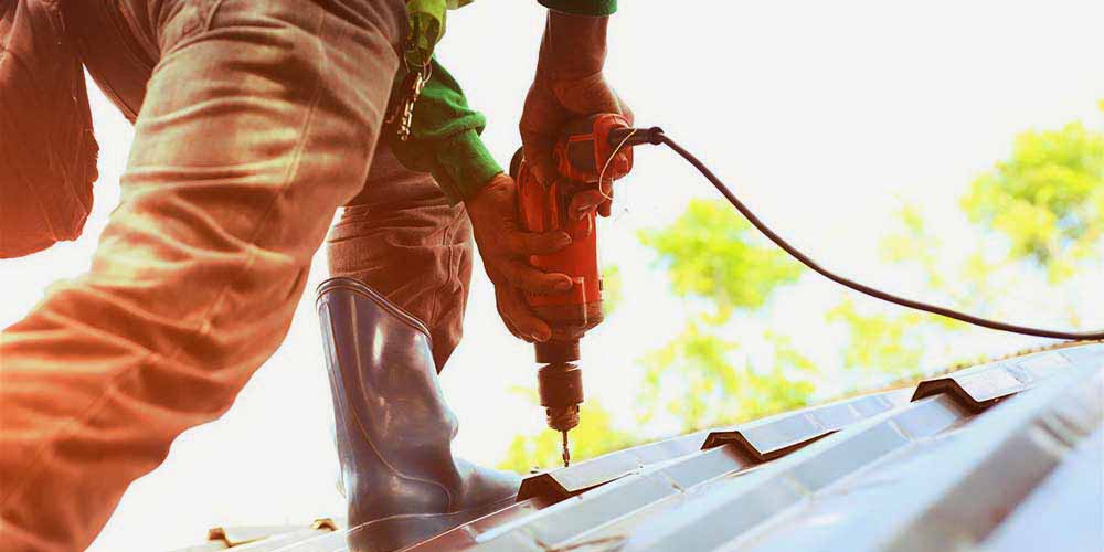 A worker using a red electric drill on a corrugated metal roof.