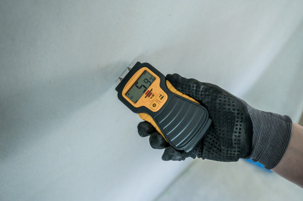 Gloved hand using a digital moisture meter on a white wall, with the display showing 5.9%.
