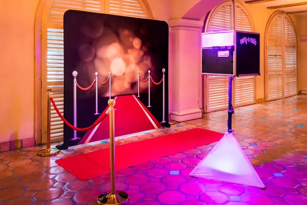 Red carpet photo booth setup with a themed backdrop, gold stanchions, and pink ambient lighting.