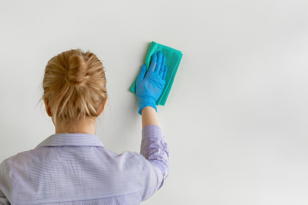 A woman wearing a blue glove and a striped shirt cleans a plain white wall with a green cloth.