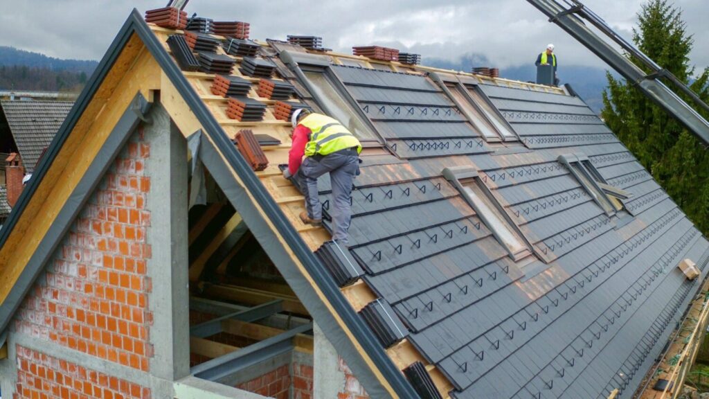 Construction workers in safety vests installing tiles on a house roof under construction.