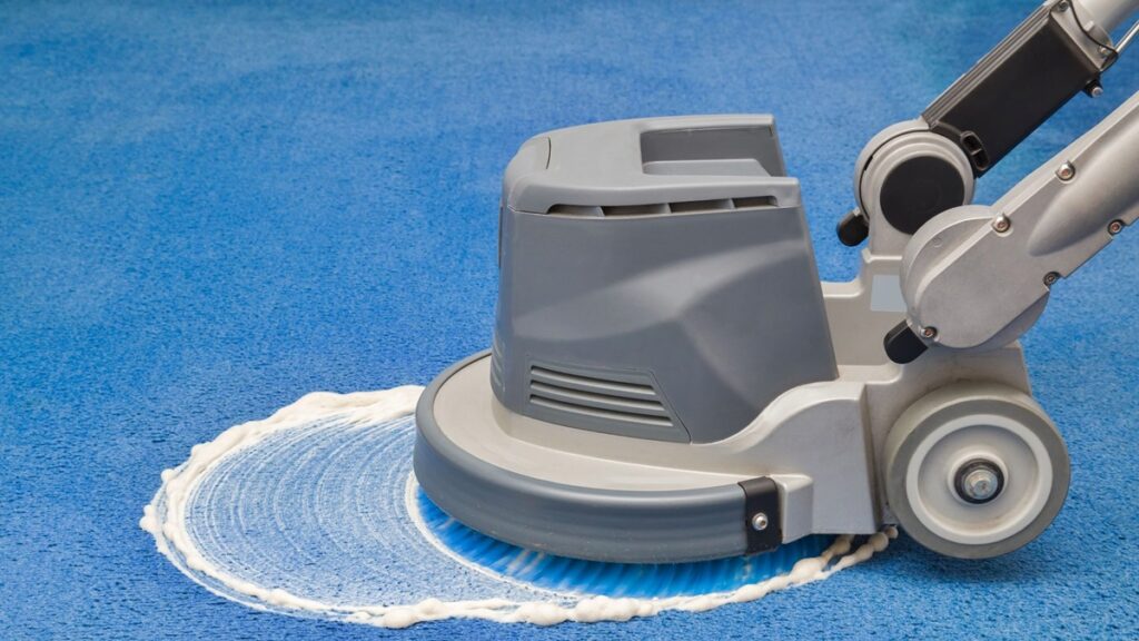 A gray floor scrubber machine cleaning a blue surface with white soapy foam.