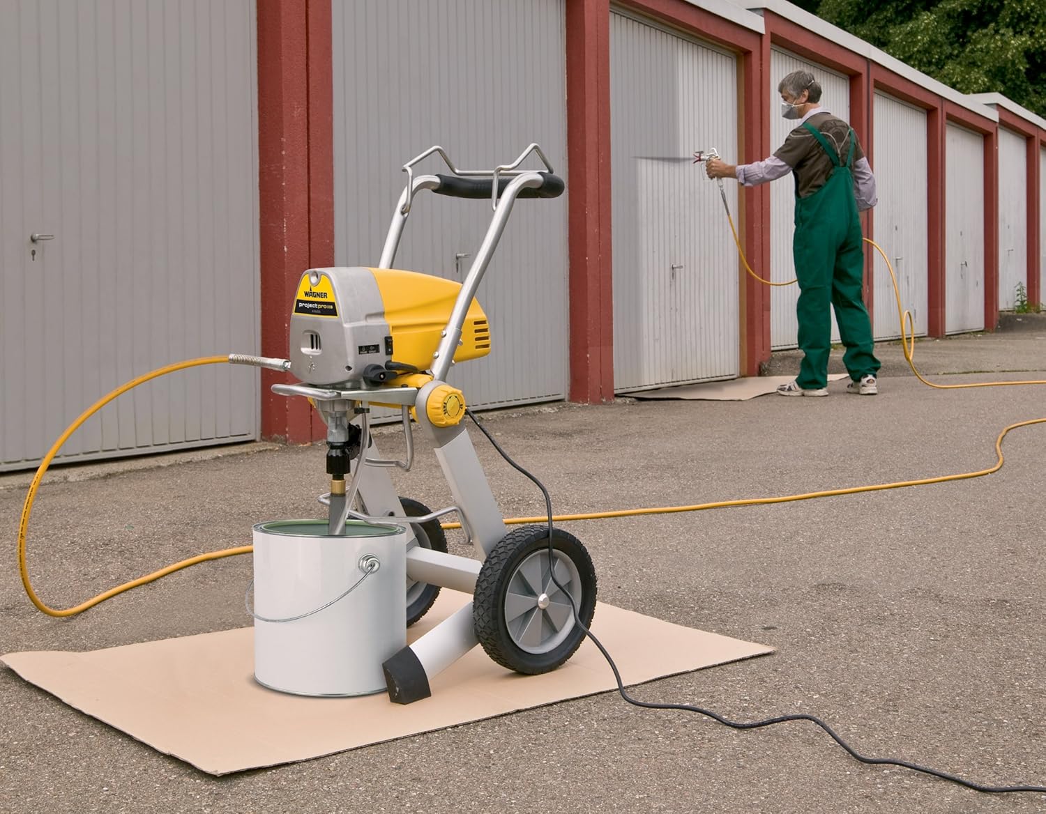 A person in green overalls uses a professional Wagner paint sprayer to coat a garage door.