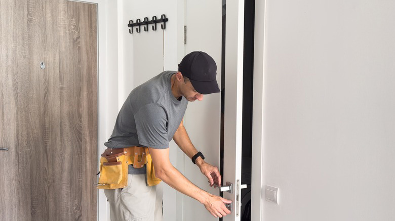 Handyman in a tool belt installing or adjusting a metal door handle on a white door.