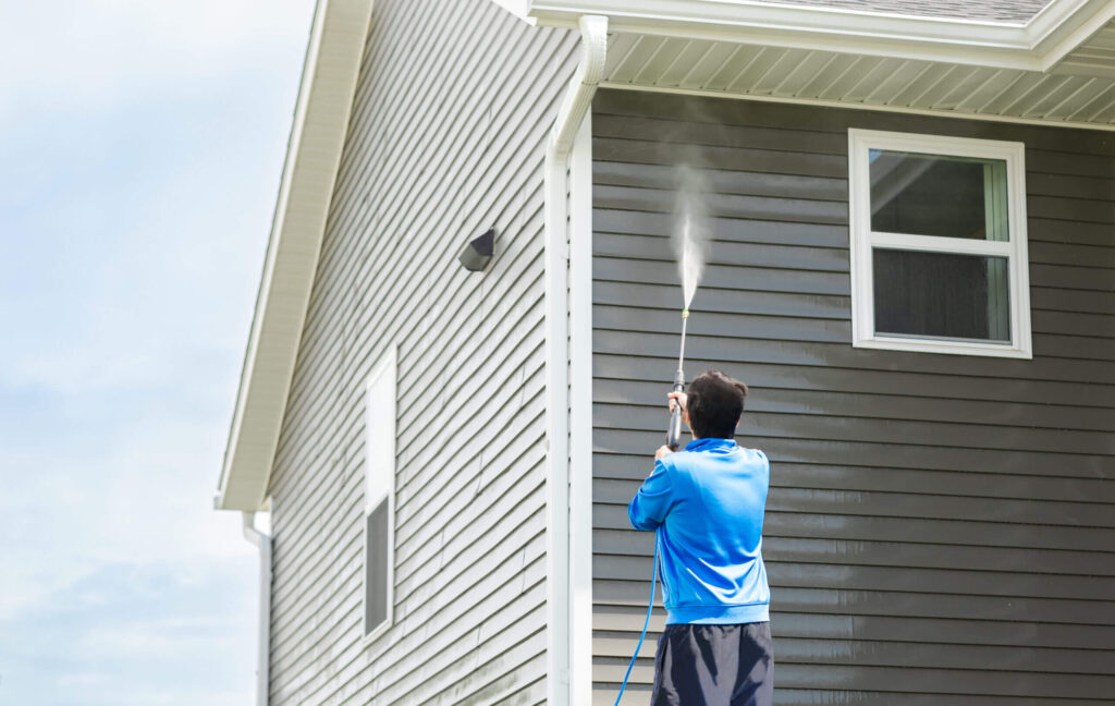 Person in a blue jacket pressure washing the grey siding of a house exterior.