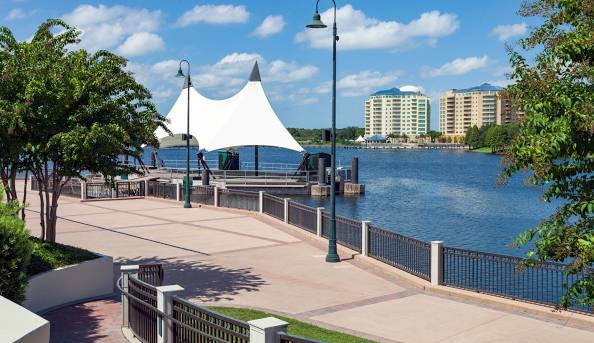Waterfront promenade with a large white canopy, paved walkway, and high-rise buildings across the water.