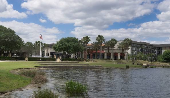 Modern apartment and retail complex with palm trees, a grassy lawn, and a pond in the foreground.