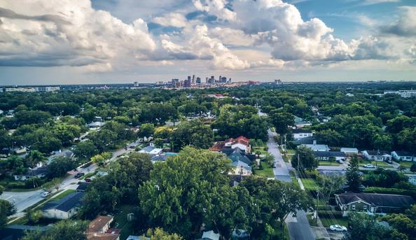 Aerial view of a leafy suburb with a distant city skyline under a dramatic, cloudy sky.