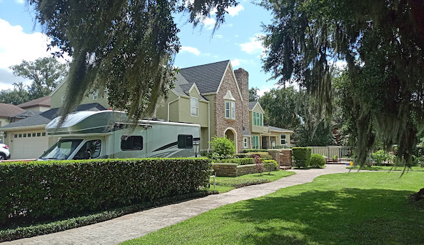 White motorhome parked in front of a large tan and stone house, framed by green hedges and trees with Spanish moss.