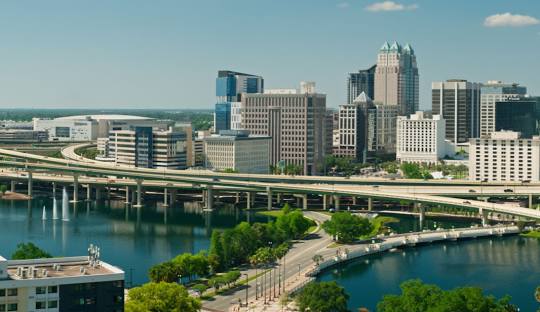Skyline of a city with modern skyscrapers, a winding highway over water, and a riverfront park with green trees.