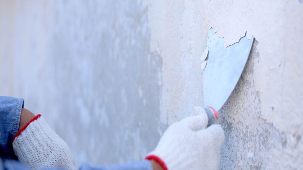 A person wearing white gloves uses a metal putty knife to scrape peeling paint off a concrete wall.