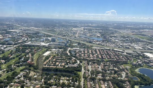 Aerial view of a sprawling suburban landscape with residential neighborhoods, highways, and city buildings under a blue sky.