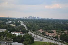 High-angle view of a road passing through a leafy suburb toward a distant city skyline under a cloudy sky.
