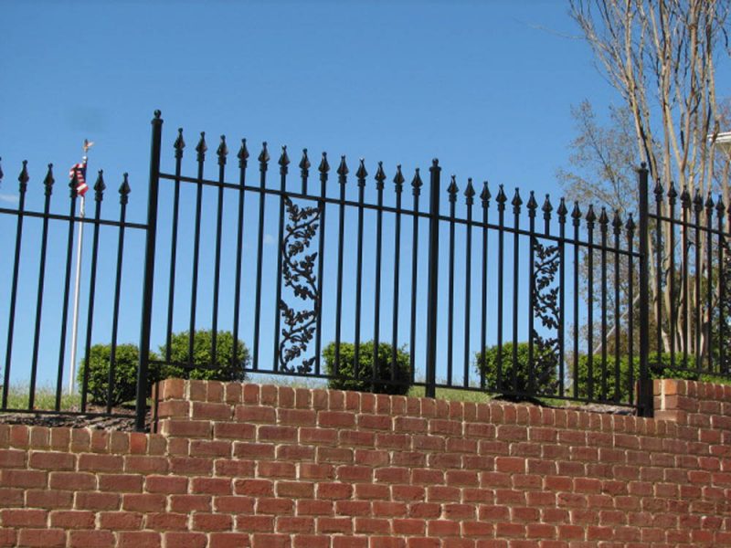 Black wrought iron fence with decorative leaf accents on top of a red brick retaining wall against a blue sky.